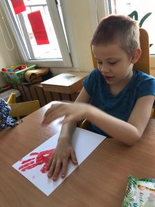 Chłopiec siedzący przy stoliku, odbijający pomalowaną czerwoną farbą dłoń na kartce papieru.