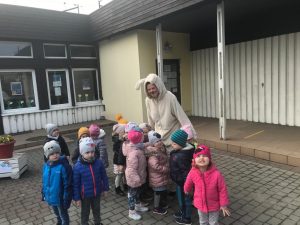 Grupa dzieci stojąca w ogrodzie przedszkolnym wraz z Panią ubraną w strój zajączka wielkanocnego.