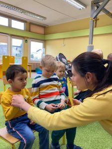 Dzieci siedzą na krzesełkach. Nauczycielka pokazuje żółtą kartkę, a dziecko wskazuje ten kolor na swojej bluzce.