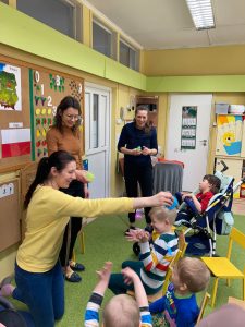 3 nauczycielki stoją przed dziećmi, które siedzą na krzesełkach i wózkach. 2 Panie leją dzieci wodą, a jedna posypuje dziecko papierowymi kroplami deszczu