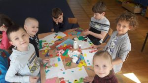 Grupa dzieci siedzi przy stoliku. Przed nimi leżą kartki z wykonaną pracą plastyczną, klej w słoiczku, kolorowe karteczki.
