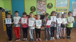Grupa dzieci stoi z rzędzie. Każde dziecko trzyma kartkę z różną literą. Powstał napis : Dzień Ziemi.