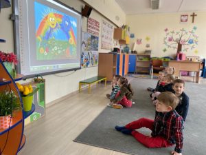 Grupa dzieci siedzących na dywanie i oglądajaca bajkę na tablicy multimedialnej.