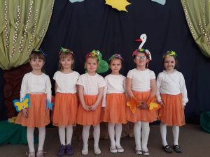Sześcioro dziewczynek stojacych w jednym rzędzie, ubranych w białe buzki i pomarańczowe spódniczki. Na głowach mają wiosenne opaski. W tle znajduje się ilustracja bociana, żaby, drzewa, słońca oraz chmurek.