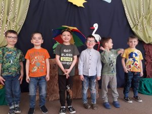 Sześcioro chłopców stojących w jednym rzędzie. Chłopiec stojący po środku ma nad głową parasol. W tle widać ilustrację bociana, drzew i słoneczka.