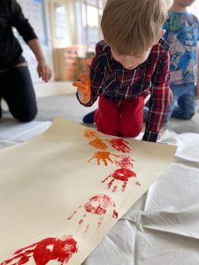 Chłopiec siedzacy na dywanie i odbijający pomalowaną na pomarańczowo dłoń na brystolu. W tle, po lewej stronie klęczy Pani, po prawej stronie chłopiec.