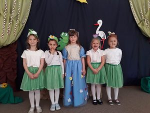 Pięć dziewczynek stojących w jednym rzędzie. Dziewczynka stojąca po środku przebrana jest za Panią Wiosnę, Dziewczynki stojące po bokach mają ubrane białe bluzki i zielone spódniczki. Na głlowach mają założone wiosenne opaski.