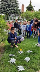 Dzieci stoją w grupie na trawie. Przed nimi rozrzucone są obrazki pająków. Jedno dziecko trzyma w ręce łapkę na muchy i schyla się do obrazków. Dziecko z łapką jest trzymane przez Pania.