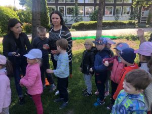 Grupa dzieci wraz z dwoma Paniami stojaca w ogrodzie przedszkolnym. Jedna Pani trzyma za rękę dwóch chłopców, druga robi pieczątki na rękach dzieci.