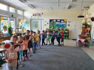 Grupa dzieci ustawionych w pociag i idących po dywanie w sali przedszkolnej. Chłopiec prowadzący pociąg trzyma w reku lizaka kolejowego.