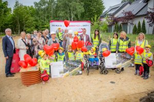 Zdjęcie grupowe przedszkolaków ubranych w kaski i kamizelki odblaskowe wraz z osobami dorosłymi. Dzieci trzymają w rękach czerwone balony, Dwoje dzieci siedzi w wózkach. Cztery Panie stojace po prawej stronie mają założone kamizelki odblaskowe. Zdjęcie wykonane jest na dworze.
