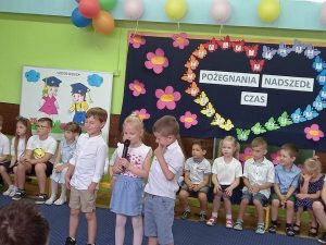Duża grupa dzieci siedzi na ławce w jednym rzędzie. Przed nimi stoi dziewczynka i 2 chłopców. Dziewczynka ma w ręce mikrofon.