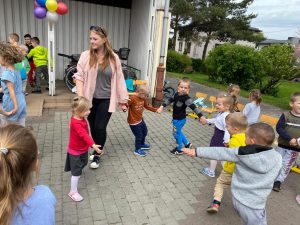 Grupa dzieci tańcząca z Panią w kole na placu przedszkola. Po lewej stronie widocznych jest tańczących sześcioro dzieci.