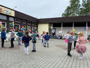 Grupa dzieci tańczących w parach przed budynkiem przedszkola, który jest udekorowany kolorowymi balonami i wiatrakami.