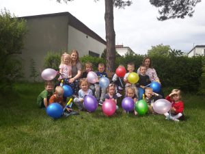 Grupa dzieci pozujaca do zdjęcia w ogrodzie przedszkolnym i trzymajaca w rękach kolorowe balony. Z dziećmi siedzą dwie Panie.