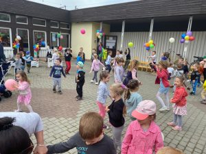 Dzieci tańczące na placu przed przedszkolem, który udekorowany jest balonami i wiatrakami.