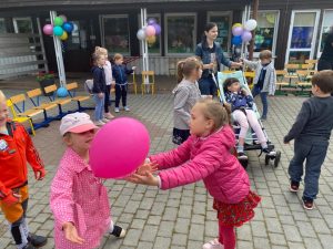 Dwie dziewcznki łapiące różowy balon na placu przedszkola. W tle widać dzieci oraz Panią trzymającą wózek, w którym siedzi dziewczynka.