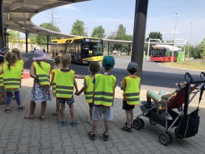 Dziesięcioro dzieci znajdujących się na placu centrum przesiadkowego i oglądajacych odjeżdzające autobusy. Chłopiec po prawej stronie siedzi w wózku.