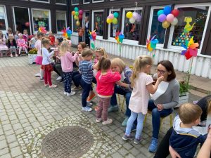 Dzieci karmiące Panie siedzące na krzesłach przed przedszkolem. Po lewej stronie, na krzesłach siedzą dzieci. Budynek przedszkola udekorowany jest balonami i wiatrakami.