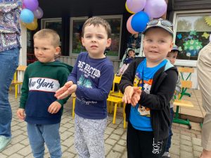 Trzech chłopców stojących przed przedszkolem. W tle widać dziecko siedzące w wózku, który trzyma Pani.