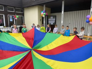 Trzynaścioro dzieci znajdujących się przedz przedszkolem i trzymających w rękach kolorową chustę. Budynek przedszkola jest udekorowany balonami.