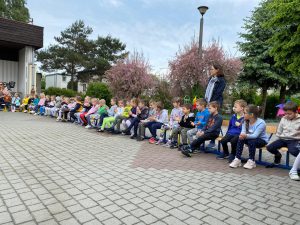 Dzieci siedzace w ogrodzie przedszkolnym na krzesłach w jednym rzędzie. Za dziećmi stoi Pani.