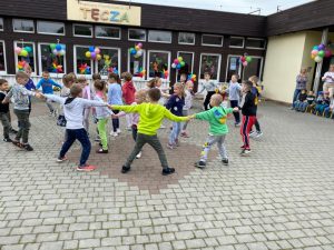 Grupa dzieci tańczących w kołach na placu przedszkolnym. W tle, po prawej stronie widoczne są dwie Panie nauczycielki. Budynek przedszkola udekorowany jest balonami oraz wiatrakami.