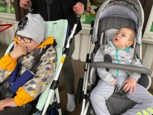 Dwóch chłopców siedzących we wózkach. Jeden wózek jest trzymany przez Panią.