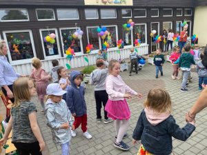 Dzieci tańczące na placu przedszkolnym. W tle widoczne są cztery Panie. Budynek przedszkola jest udekorowany kolorowymi balonami i wiatrakami.