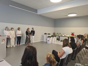 Duża sala w której na krzesłach siedzą Panie w maseczkach. 4 Panie stoją pod ścianą.