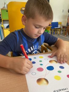 Chłopiec siedzi przy stoliku.Łączy pisakiem na kartce kolorowe kropki.