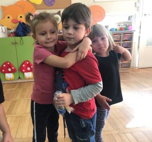 Dziewczynka przytulająca chłopca, który trzyma w ręku upominek. Z tyłu stoi dziewczynka.