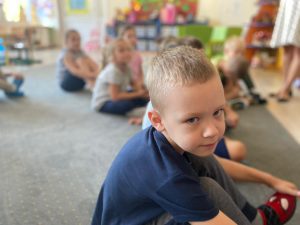 Chłopiec siedzący na dywanie w sali przedszkolnej. W tle widoczna grupa dzieci.