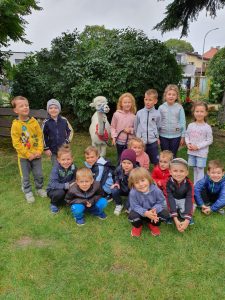 Grupa dzieci pozująca do zdjęcia w ogrodzie przedszkolnym. Pomiędzy dziećmi, w drugim rzędzie stoi alpaka, którą trzyma dziewczynka.
