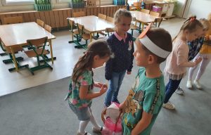 Na pierwszym tle troje dzieci stojących w sali przedszkolnej. Chłopiec częstuje dziewczynki lizakami. W tle stoją trzy dziewczynki.
