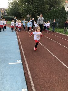 Chłopiec biegnący na bieżni sportowej. Za nią stoi grupa dzieci oraz trzy osoby dorosłe.