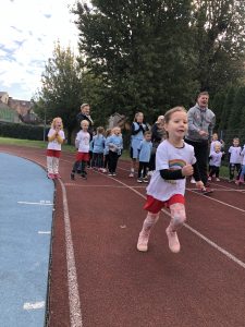 Dziewczynka biegnąca na bieżni sportowej. Za nią stoi grupa dzieci oraz trzy osoby dorosłe.