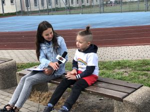 Chłopiec z nastolatką siedzący na ławce, na boisku szkolnym. Dziewczyna trzyma w ręku mikrofon, chłopiec udziela wywiadu.