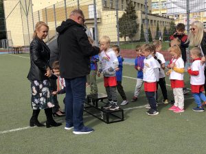 Grupa dzieci czekająca na wręczenie medali na boisku sportowym. Przed dziećmi stoi mężczyzna i kobieta, Za dziećmi stoi kobieta i kamerzysta.