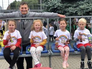 Czworo dzieci siedzących na trybunach obok siebie, jedzących wafelki i trzymających napoje. Za dziećmi stoi mężczyzna z rozłożonymi rękami.