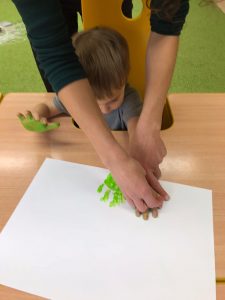 Chłopiec siedzi przy stole ma pomalowane dłonie na zielono, które odciska na kartce papieru.