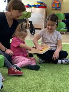 2 dziewczynki i Pani. Siedzą na dywanie i dotykają makaron na tacy.