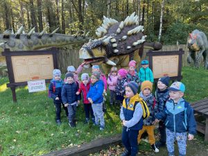 Grupa dzieci pozuje do zdjęcia przy figurze dinozaura. Widoczne są również tablice informacyjne o znajdujących się tam dinozaurach.