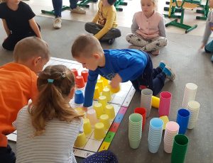 Troje dzieci pochyla się nad rozłożoną matą, z kolorowymi kubeczkami.