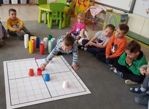 Na dywanie znajduje się kwadratowa mata, na której chłopiec układa kolorowe kubki według określonego wzoru. Obok siedzą dzieci i obserwują co robi kolega.