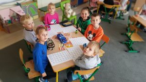 Sześcioro dzieci siedzi przy stoliku, na którym znajdują się piórniki oraz karty pracy , przygotowane do wykonania zadania.