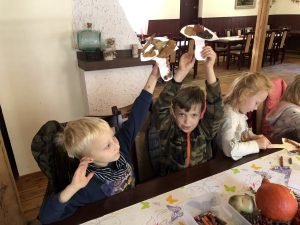 Czworo dzieci siedzących obok siebie przy stole. Dwóch chłopców trzyma ręce do góry z papierowymi grzybkami. Jedna dziewczynka koloruje papierowego grzybka