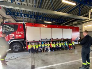 Z prawej strony stoi strażak czytający dzieciom bajkę. Z lewej strony także stoi strażak. Po środku grupa dzieci siedzących przed wozem strażackim.