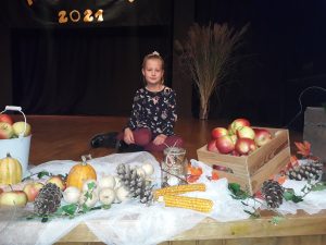 Dziewczynka siedzi bokiem a scenie otoczona dekoracją z liści jabłek, szyszek oraz dyni.