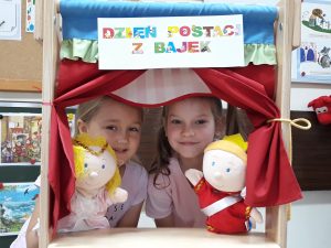 Dwie dziewczynki z pacynkami Jasia i Małgosi stoją za mini sceną . Nad głowami dziewczynek widnieje napis Dzień Postaci z Bajek.